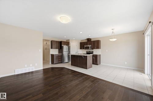 Open-concept living area featuring dark wood-finish flooring, a kitchen with dark cabinetry, stainless steel appliances, a center island with a sink, and light-toned tile flooring - 2170 52A Street Sw, Edmonton, AB - Indoor Photo Showing Kitchen
