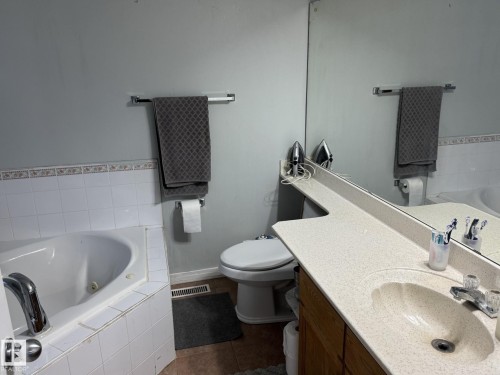 Bathroom featuring a built-in tub with white tile surround, a vanity with an integrated sink and light-toned countertop, a large wall-mounted mirror, and a toilet - 3139 34B Avenue, Edmonton, AB 