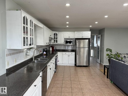 Kitchen featuring white cabinetry with glass-front uppers, dark countertops, a stainless steel double sink with a gooseneck faucet, and tile flooring - 3139 34B Avenue, Edmonton, AB 
