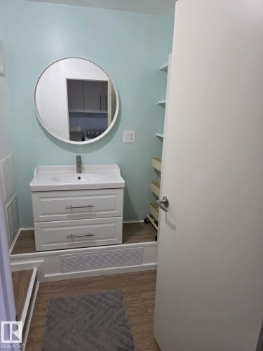 White vanity with integrated sink and chrome faucet, round mirror, and wood-finish flooring - 4825 52 Street, Elk Point, AB - Indoor Photo Showing Bathroom