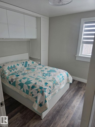 Integrated white cabinetry and shelving above and beside the bed, complemented by a window with blinds, wood-finish flooring, and a ceiling-mounted light fixture. - 4825 52 Street, Elk Point, AB - Indoor Photo Showing Bedroom