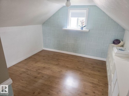 Room featuring a window, sloped ceiling, wood-finish flooring, light blue patterned accent wall, and white baseboards - 4825 52 Street, Elk Point, AB - Indoor Photo Showing Other Room