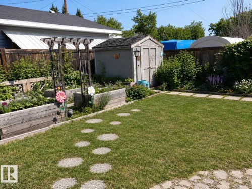 Expansive backyard featuring a manicured lawn, a stepping stone path, raised garden beds with a trellis, a storage shed, and perimeter fencing. - 4825 52 Street, Elk Point, AB - Outdoor