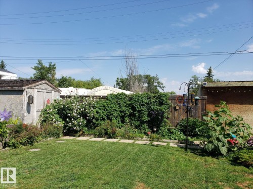 Expansive backyard featuring a green lawn, stepping stones, garden beds with various plantings, a wooden storage shed, and a privacy fence. - 4825 52 Street, Elk Point, AB - Outdoor