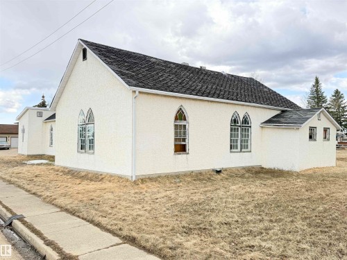 White stucco exterior featuring a gabled roofline with dark shingles, multiple arched windows, and white gutters - 5036 49 Street, Clyde, AB - Outdoor