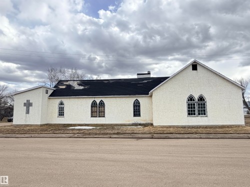White textured stucco exterior with Gothic-style arched windows, a black asphalt shingle roof, and a prominent cross architectural element - 5036 49 Street, Clyde, AB - Outdoor