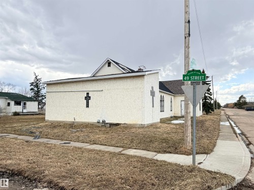 Stucco exterior building with a prominent cross architectural detail, multiple arched windows, and a black asphalt shingle roof - 5036 49 Street, Clyde, AB - Outdoor