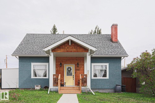 Exterior featuring a blue stucco finish, a covered front porch with wood shingle siding, a yellow front door, a brick chimney, and a gray shingle roof - 5113 53 Avenue, Stony Plain, AB - Outdoor
