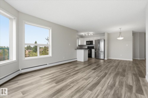 Open-concept living area featuring wood-finish flooring, large windows, recessed lighting, and a modern kitchen with stainless steel appliances - 407 13635 34 Street Nw, Edmonton, AB - Indoor Photo Showing Other Room