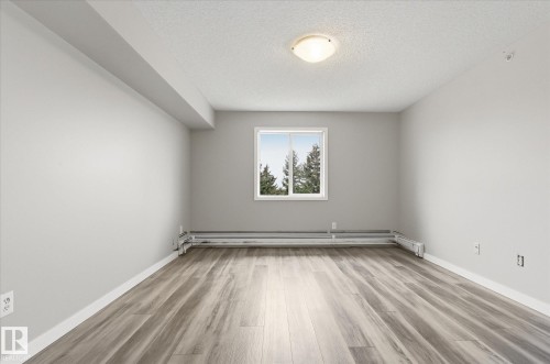 Spacious room featuring wood-finish flooring, a white window frame, and baseboard heating - 407 13635 34 Street Nw, Edmonton, AB - Indoor Photo Showing Other Room
