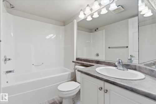 Full bathroom featuring a white tub and shower combination with chrome fixtures, a white toilet, and a vanity with white cabinetry, a gray countertop, and an integrated sink with a chrome faucet - 407 13635 34 Street Nw, Edmonton, AB - Indoor Photo Showing Bathroom