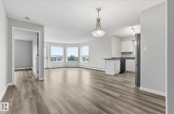Open-concept living area with wood-finish flooring, a bay window offering exterior scenery, and a kitchen featuring white cabinetry and black appliances - 