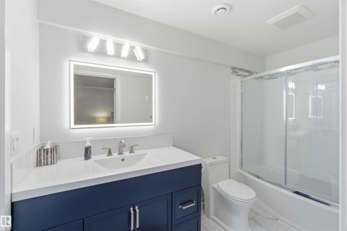 Bathroom featuring a vanity with a white countertop and blue cabinetry, an illuminated mirror, and a shower with a sliding glass door - #1 Colonial Close, Beaumont, AB - Indoor Photo Showing Bathroom