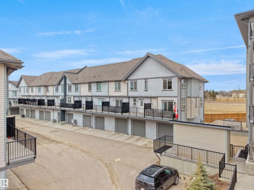 The property is a multi-story townhome featuring a private balcony with black railings, a garage with a roll-up door, and a light-colored exterior with white trim - 64 655 Watt Boulevard, Edmonton, AB - Outdoor With Exterior