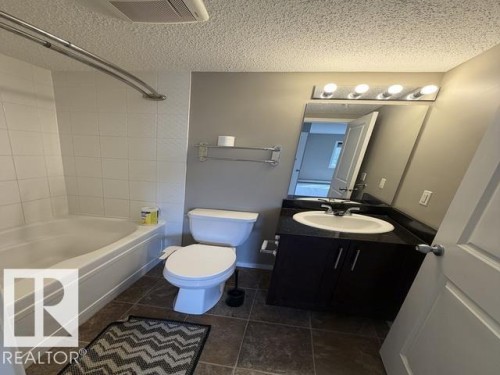 Bathroom featuring a white bathtub with subway tile surround, a dark wood-finish vanity with an integrated sink, a dark countertop, and a chrome light fixture with four bulbs - 321 107 Watt Common, Edmonton, AB - Indoor Photo Showing Bathroom