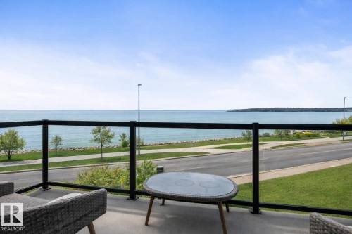 The property features a balcony with a glass railing, providing views of the water, a paved road, and a landscaped green area with trees - 515 Lakeshore Drive, Cold Lake, AB - Outdoor With Body Of Water With View
