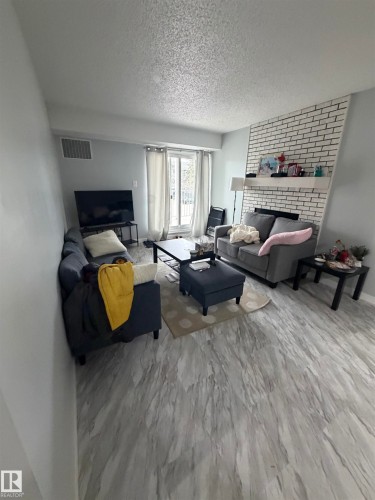 Living area featuring light-toned wood-finish flooring, a white brick-patterned fireplace with a mantel, and a sliding glass door - 205 10136 160 Street, Edmonton, AB - Indoor Photo Showing Living Room With Fireplace