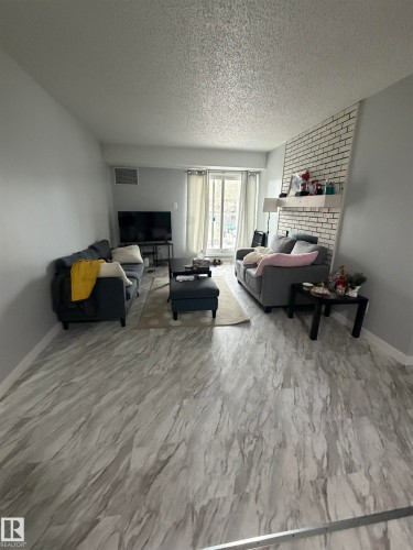 Spacious living area featuring light-toned wood-finish flooring and a white painted brick accent wall - 205 10136 160 Street, Edmonton, AB - Indoor Photo Showing Living Room