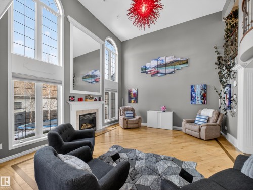 Grand living space featuring expansive arched windows, a built-in fireplace with a white mantel, high ceilings, wood-finish flooring, and a decorative staircase - 1062 Tory Road, Edmonton, AB - Indoor Photo Showing Living Room With Fireplace