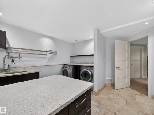 Laundry room featuring a large central island with a light-colored countertop, dark wood-finish cabinetry, a built-in sink with a gooseneck faucet, two front-load laundry appliances, and light-toned tile flooring - 1062 Tory Road, Edmonton, AB - Indoor