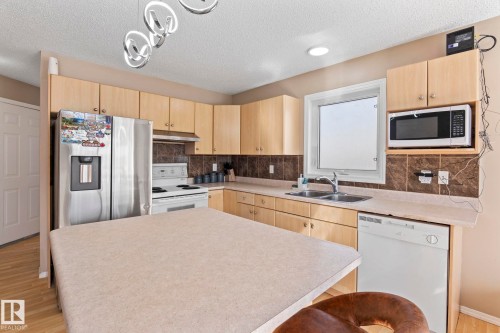 Kitchen featuring light wood-finish cabinetry, a central island with countertop, stainless steel refrigerator, white range, and a dual basin sink beneath a window - 92 4350 23 Street, Edmonton, AB - Indoor Photo Showing Kitchen With Double Sink