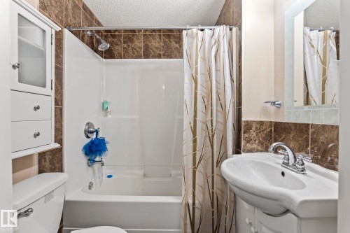 Bathroom featuring a white bathtub with an integrated shower, chrome fixtures, a wall-mounted sink with a chrome faucet, and a wall cabinet with glass-fronted storage - 92 4350 23 Street, Edmonton, AB - Indoor Photo Showing Bathroom