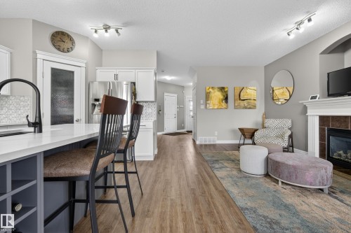 Open-concept living area featuring wood-finish flooring, a kitchen island with built-in storage, stainless steel appliances, a tiled fireplace with a white mantel, and track lighting - 2146 Austin Link Link, Edmonton, AB - Indoor Photo Showing Living Room With Fireplace