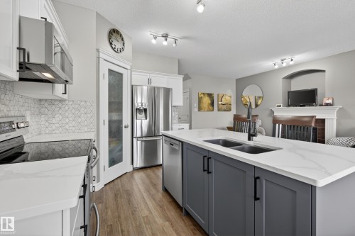 Contemporary kitchen featuring an expansive island with a dual-basin sink and matte black faucet, wood-finish flooring, white cabinetry, geometric tile backsplash, and stainless steel appliances - 2146 Austin Link Link, Edmonton, AB - Indoor Photo Showing Kitchen With Double Sink With Upgraded Kitchen