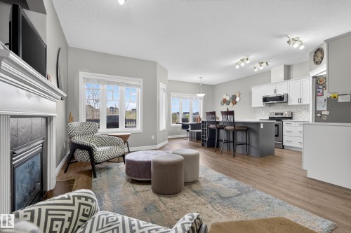 Open-concept living area featuring wood-finish flooring, a white fireplace surround with dark tile, a kitchen island with seating, white cabinetry, and stainless steel appliances - 2146 Austin Link Link, Edmonton, AB - Indoor With Fireplace