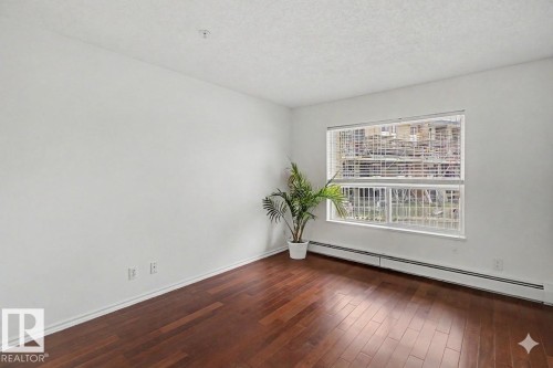 Spacious room featuring rich wood-finish flooring, white baseboards, and a large window with horizontal blinds - 213 17404 64 Avenue, Edmonton, AB 