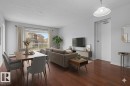 Open-concept living space with dark wood-finish flooring, light-toned walls, and a large window - 213 17404 64 Avenue, Edmonton, AB 