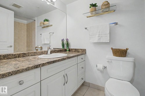 Bathroom featuring a white vanity with paneling and brushed nickel hardware, a speckled countertop, an undermount sink with a chrome faucet, a large wall-mounted mirror, and tile flooring - 213 17404 64 Avenue, Edmonton, AB 