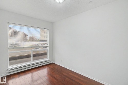 Bright room featuring a large window with blinds, wood-finish flooring, white walls, and baseboard heating - 213 17404 64 Avenue, Edmonton, AB 