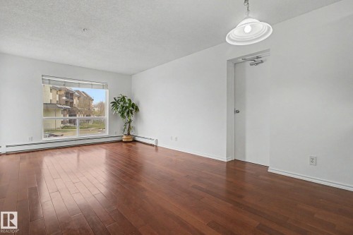Spacious interior featuring wood-finish flooring, a large window, white walls, and a ceiling-mounted light fixture - 213 17404 64 Avenue, Edmonton, AB 