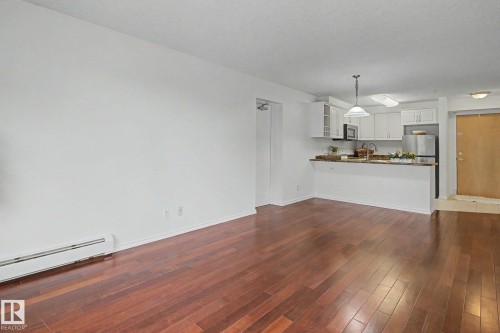 Open-concept living space featuring rich wood-finish flooring, white walls, and a kitchen area with white cabinetry, stainless steel appliances, and a peninsula countertop - 213 17404 64 Avenue, Edmonton, AB 