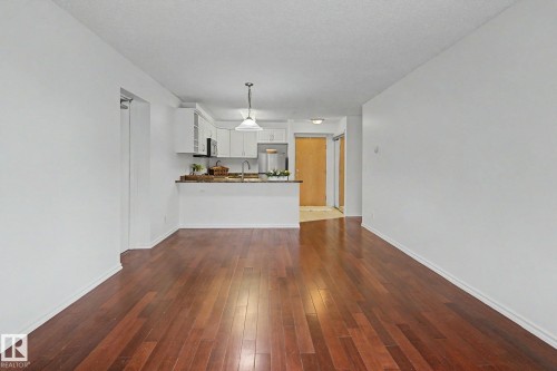 Hardwood flooring throughout a bright living area, featuring a kitchen with white cabinetry, light-toned countertops, and a stainless steel refrigerator - 213 17404 64 Avenue, Edmonton, AB 