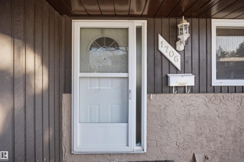 Front entry with a white storm door, vertical wood panel siding, decorative outdoor lighting, and a textured stucco foundation - 1706 37 Street, Edmonton, AB - Outdoor With Exterior