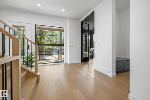 Entryway featuring light wood-finish flooring, a contemporary staircase with wood handrails and black metal spindles, a spacious window with black framing, a white entry door with glass inserts, and a built-in storage bench with dark cabinetry - 13804 85 Avenue, Edmonton, AB - Indoor Photo Showing Other Room