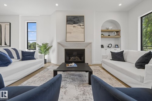 Spacious interior featuring light wood-finish flooring, a contemporary fireplace with a stone-finish mantle, large windows, and a built-in arched alcove with shelving and lower cabinetry - 13804 85 Avenue, Edmonton, AB - Indoor Photo Showing Living Room With Fireplace