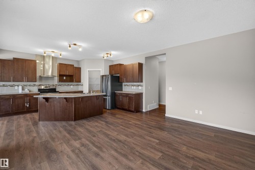 Kitchen featuring wood-finish flooring, dark wood cabinetry, a central island with integrated sink, stainless steel appliances, and a tiled backsplash - 4723 Alwood Bend Sw, Edmonton, AB - Indoor Photo Showing Kitchen