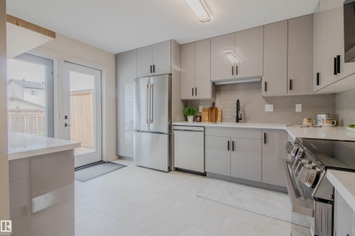 Modern kitchen featuring sleek gray cabinetry, stainless steel appliances, light-toned countertops, a full-height tile backsplash, and a glass-paned door leading to the exterior - 5935 189 Street, Edmonton, AB - Indoor Photo Showing Kitchen
