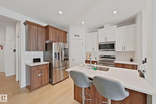 Contemporary kitchen featuring a large island with seating, stainless steel appliances, a white mosaic tile backsplash, and two-tone cabinetry with wood-finish and white upper cabinets - 1231 Watt Drive, Edmonton, AB - Indoor