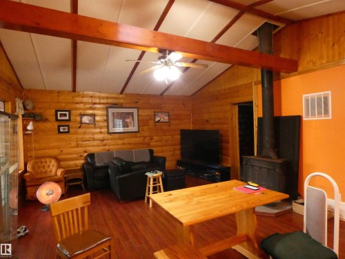Vaulted ceiling with exposed wood beams, log-style wall paneling, rich wood-finish flooring, and a freestanding wood-burning stove with a black stovepipe - 307 Willow Place, Rural Barrhead County, AB 