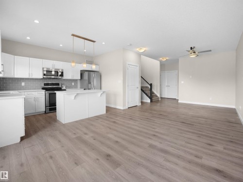 Open-concept kitchen and living area featuring wood-finish flooring, white cabinetry, stainless steel appliances, a kitchen island, and a staircase with dark banisters - 8921 217 Street Nw, Edmonton, AB - Indoor Photo Showing Kitchen