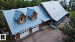 Property exterior featuring a brown siding finish, light blue metal roofing, two prominent dormers, two white garage doors, and an extended covered porch - 