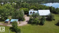 Expansive greenspace surrounding a property featuring a main residence with dormer windows, an additional structure with a light blue roof, and a detached shed, all situated near a body of water - 