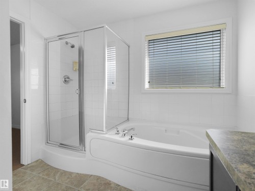 Bathroom featuring a white built-in bathtub with deck-mounted fixtures, an adjacent glass-enclosed shower, and a window with horizontal blinds - 2017 32A Street, Edmonton, AB - Indoor Photo Showing Bathroom