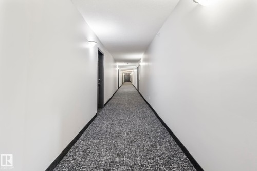 Hallway featuring wall-mounted sconce lighting, white walls, dark baseboards, and patterned carpet flooring - 412 5005 165 Avenue Nw, Edmonton, AB - Indoor Photo Showing Other Room