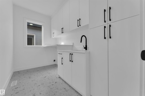 Laundry area featuring white cabinetry with black hardware, a utility sink with a matte black faucet, a window with white trim, and patterned tile flooring - 12 Dalquist Bay, Leduc, AB - Indoor