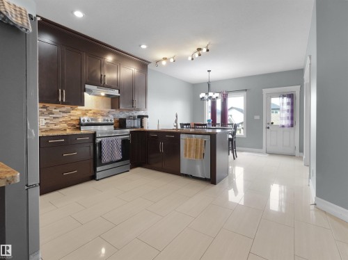 Open-concept kitchen featuring dark wood cabinetry, stainless steel appliances, a multi-tone tile backsplash, and a central island with an integrated sink - 5192 1B Ave Sw, Edmonton, AB - Indoor Photo Showing Kitchen